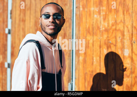 Young black man wearing casual clothes and sunglasses, smiling against a wooden background. Millennial african guy with bib pants outdoors Stock Photo