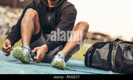 Athlete wearing sports shoes getting ready for training. Cropped shot of a runner sitting on a running track tying shoe lace with a bag by his side. Stock Photo