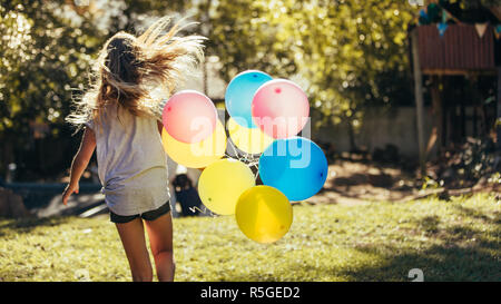 Rear view of small girl running away with colorful balloons outdoors in the backyard. Girl having fun outdoors with balloons. Stock Photo