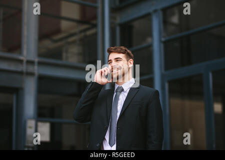 Smiling businessman talking over mobile phone while walking outside office building. Man in formal clothes walking outdoors and talking on phone. Stock Photo