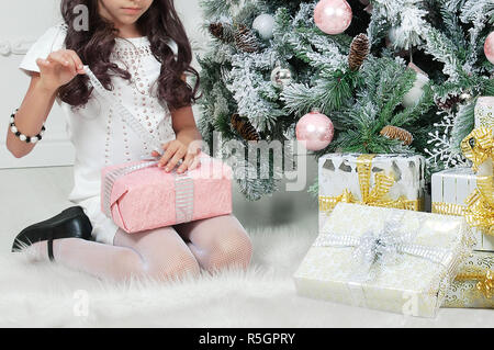 Little girl on the floor by the Christmas tree. Opens the box with a gift. Stock Photo