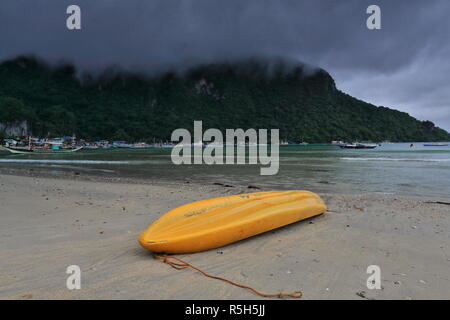 Yellow fiberglass kayak stranded upside down at the beach-tourboats moored in a rainy morning preventing them from going out to sea for fishing and to Stock Photo