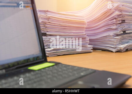 computer notebook and documents on desk stack up high waiting to be managed Stock Photo