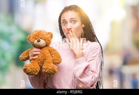Young african american girl holding teddy bear over isolated background cover mouth with hand shocked with shame for mistake, expression of fear, scar Stock Photo