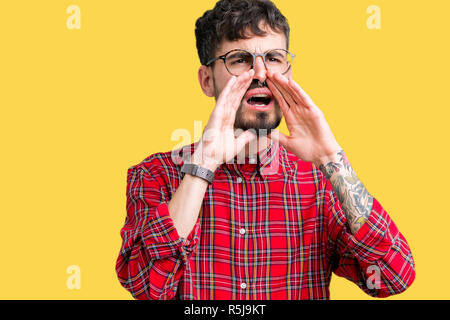 Young handsome man wearing glasses over isolated background Shouting angry out loud with hands over mouth Stock Photo