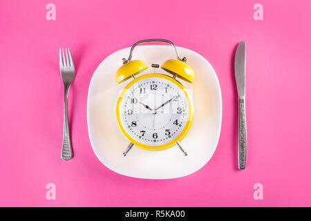 Retro alarm clock on plate with fork and knife isolated on pink background. Chrono nutrition diet concept. Top view Stock Photo