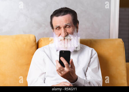 Happy senior bearded man wearing white shirt sitting on the yellow sofa and using smartphone Stock Photo