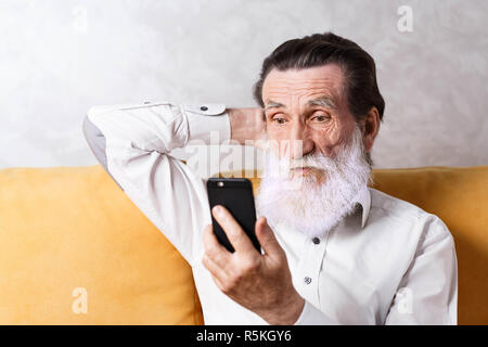 Surprised senior bearded man wearing white shirt sitting on the yellow sofa and looking on the screen of his smartphone Stock Photo