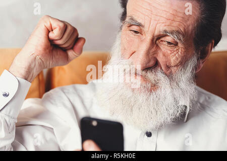 Modern senior bearded man wearing white shirt sitting on the yellow sofa and rejoicing of something on the his smartphone Stock Photo