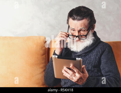 Clever senior bearded man in glasses wearing grey pullover reading electronic book on the digital tablet while sitting on a yellow sofa in his light living room, he looking over his glasses Stock Photo