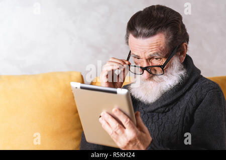 Senior bearded man in glasses wearing grey pullover sitting on a yellow sofa in his light living room and looking over his glasses and reading something on the tablet computer. Stock Photo