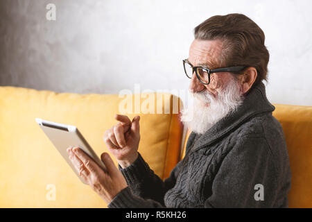 Senior bearded man in glasses wearing grey pullover sitting on a yellow sofa in his light living room and swipping facebook news on the digital tablet. Stock Photo