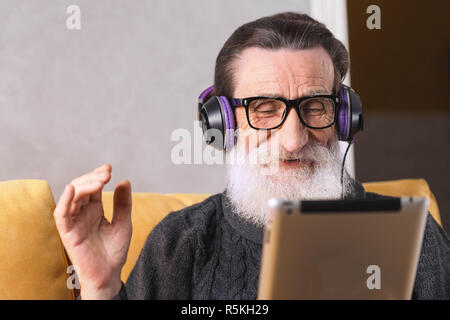 Senior cheerful bearded man in glasses wearing grey pullover sitting in headphones on a yellow sofa in his light living room and chatting with his family on Skype on the digital tablet, technology, people, lifestyle and communication concept Stock Photo