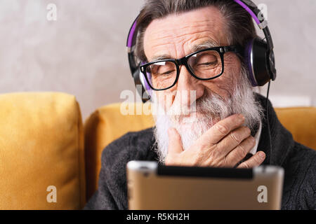 Senior thoughtful bearded man in glasses wearing grey pullover learning to use the digital tablet while sitting in headphones on a yellow sofa in his light living room, he straightens his beard Stock Photo