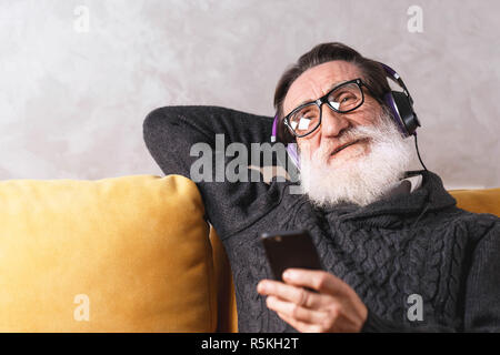 Senior cheerful bearded man in glasses wearing grey pullover relaxing with the smartphone while sitting in headphones on a yellow sofa in his light living room, modern technology, communication concept Stock Photo