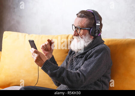 Senior cheerful bearded man in glasses wearing grey pullover using smartphone while sitting in headphones on a yellow sofa in his light living room, modern technology, communication concept Stock Photo