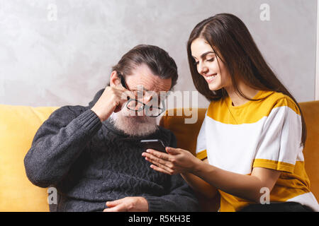 Senior handsome bearded man in glasses wearing grey pullover sitting with his adult long haired beautiful daughter on the yellow sofa and looking seriously on the smartphone, modern technology, communication concept Stock Photo