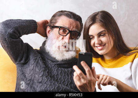 Surprised senior bearded father in glasses using smartphone while sitting on the yellow sofa in the light living room with his adult long haired brunette daughter, he shocked of the modern technologys, modern technology, communication concept Stock Photo