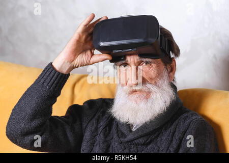 Contemporary senior bearded man wearing grey pullover sitting in VR goggles on the yellow sofa in the light living room, he looking at the camera, future technology concept Stock Photo