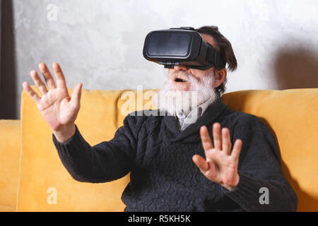 Surprised senior bearded man in grey pullover gesturing while testing VR device, he sitting on the yellow sofa in the light living room, future technology concept Stock Photo