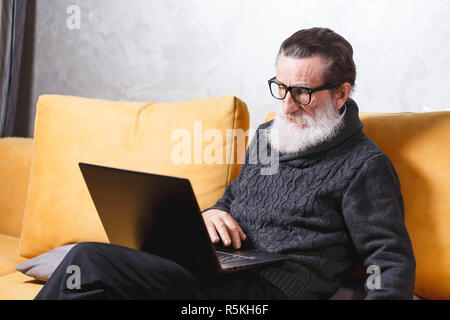 Handsome senior bearded man in glasses and grey pullover sitting with laptop on the yellow sofa in the light living room, modern technology, communication concept Stock Photo