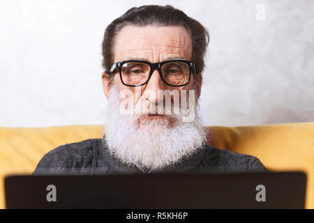 Concentrated senior bearded man in glasses and grey pullover working with laptop while sitting on the yellow sofa in the light living room, modern technology, communication concept Stock Photo