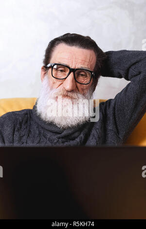 Contemporay senior bearded man in glasses and grey pullover using laptop and browsing internet while sitting on the yellow sofa in the light living room, modern technology, communication concept Stock Photo