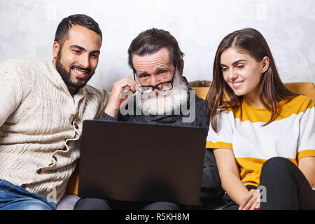 Senior bearded father in glasses sitting and watching movie with his brunette bearded son and brunette long haired daughter on the yellow sofa in the light living room, modern technology, communication concept Stock Photo