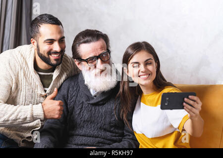 Brunette bearded son and brunette long haired daughter smiling and taking selfie with they senior bearded father in glasses while sitting on the yellow sofa in the light living room, modern technology, communication concept Stock Photo