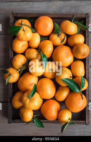Tangerines (oranges, clementines, citrus fruits) with green leaves in box on wooden background, top view. Stock Photo