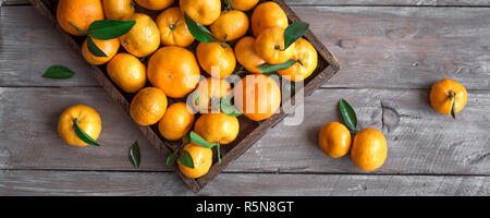 Tangerines (oranges, clementines, citrus fruits) with green leaves in box on wooden background, top view, banner. Stock Photo