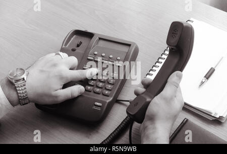 The businessman dials the number on the landline phone, holds the handset in his right hand