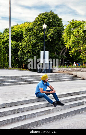 Young indian sikh sitting on the stairs of mount royal park and talking on the phone with his earphones in Montreal, Canada. Stock Photo