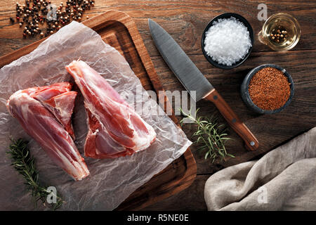 Raw lamb mutton leg drumstick on white cooking paper and wooden cutting board. Decorated with herbs, spices, chef's knife and napkin. Overhead view. Stock Photo