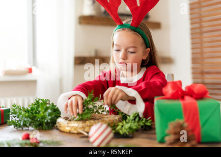 Cute preschooler girl dressed in reindeer costume wearing reindeer antlers making christmas wreath in living room. Christmas decoration family fun con Stock Photo