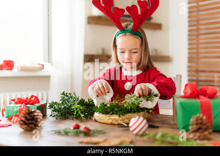 Cute preschooler girl dressed in reindeer costume wearing reindeer antlers making christmas wreath in living room. Christmas decoration family fun con Stock Photo