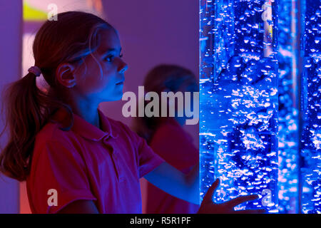 Child in therapy sensory stimulating room, snoezelen. Child interacting with colored lights bubble tube lamp during therapy session. Stock Photo