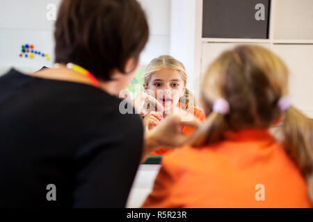 Young girl in speech therapy office. Mirror reflection of young girl exercising correct pronunciation with speech therapist. Stock Photo