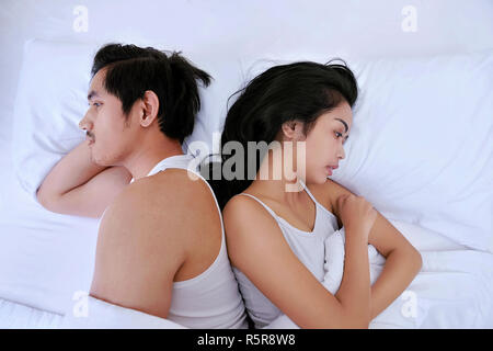 Upset asian couple having problems with lying side by side in bed facing in opposite directions