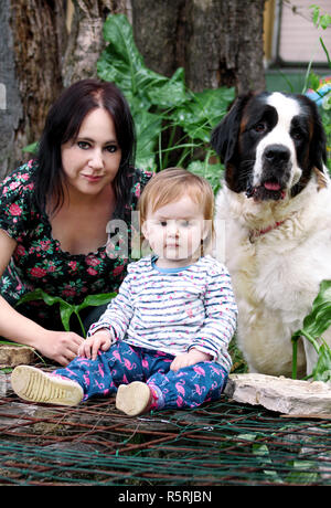 Family and love pets concept portrait with mother and baby girl is sitting and playing with dog in garden outdoors. Child and mom with St. Bernard dog Stock Photo