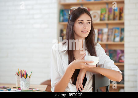 Adorable young woman posing with cup of coffee or tea in hand. Gorgeous girl looking away and sitting. Model with chestnut hair wearing in white shirt. Painter in art studio. Stock Photo