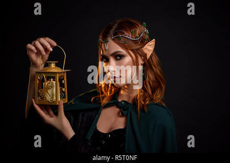 Portrait of charming mythical creature with red hair, in black dress and dark green cloak. Young beautiful elf with tiara on head holding in her hand golden lantern with white candle inside. Stock Photo