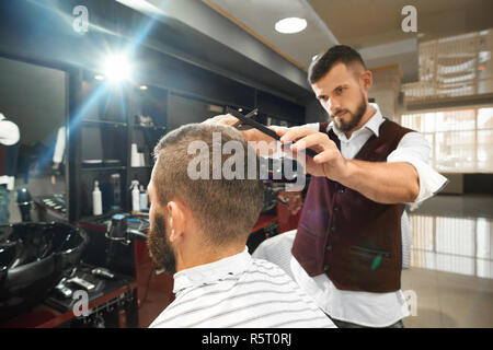 Serious brutal barber in uniform standing near male client sitting in chair and doing stylish haircut in barbershop. Qualified man using comb and straight razor.  Concept of working process. Stock Photo