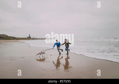 Young Couple Run Along Winter Beach With Their Dog Stock Photo