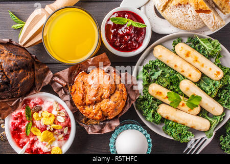 Breakfast served with muffins, grilled sausages, juice, fresh bread and parfait on a dark wooden table, top view. Stock Photo