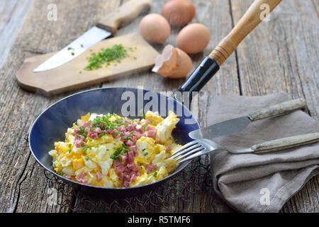 Hearty scrambled eggs with bacon and chives served steaming hot in an old enamel frying pan Stock Photo