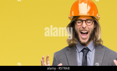 Young handsome architec man with long hair wearing safety helmet over isolated background celebrating mad and crazy for success with arms raised and c Stock Photo