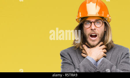 Young handsome architec man with long hair wearing safety helmet over isolated background shouting and suffocate because painful strangle. Health prob Stock Photo
