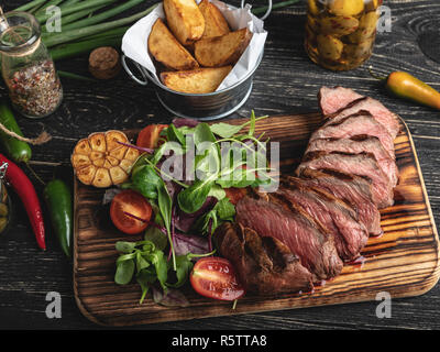 sliced juicy steak medium rare on the board with herbs, fried potatoes, spices on a black surface Stock Photo