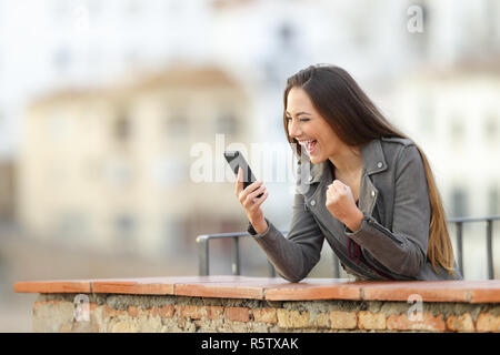 Excited woman checking smart phone in a balcony with a town in the background Stock Photo